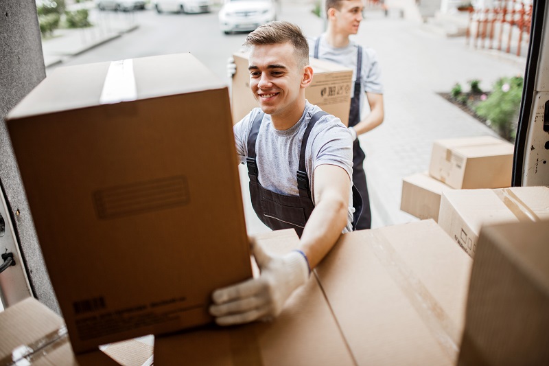 A-young-handsome-smiling-mover-wearing-uniform-is-reaching-for-the-box-while-unloading-the-van-full-of-boxes-House-move-mover-service.
